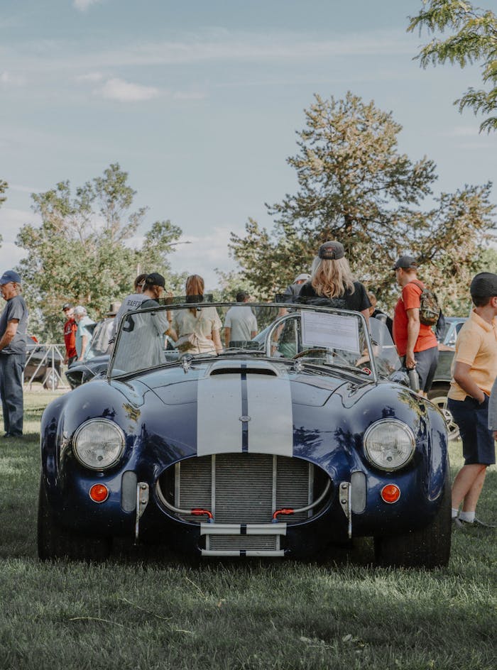 Classic blue Shelby Cobra on display at an outdoor car show with spectators.