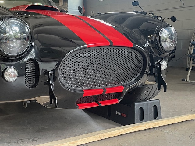 Angled front view of a black Cobra replica with red racing stripes and an installed perforated grille